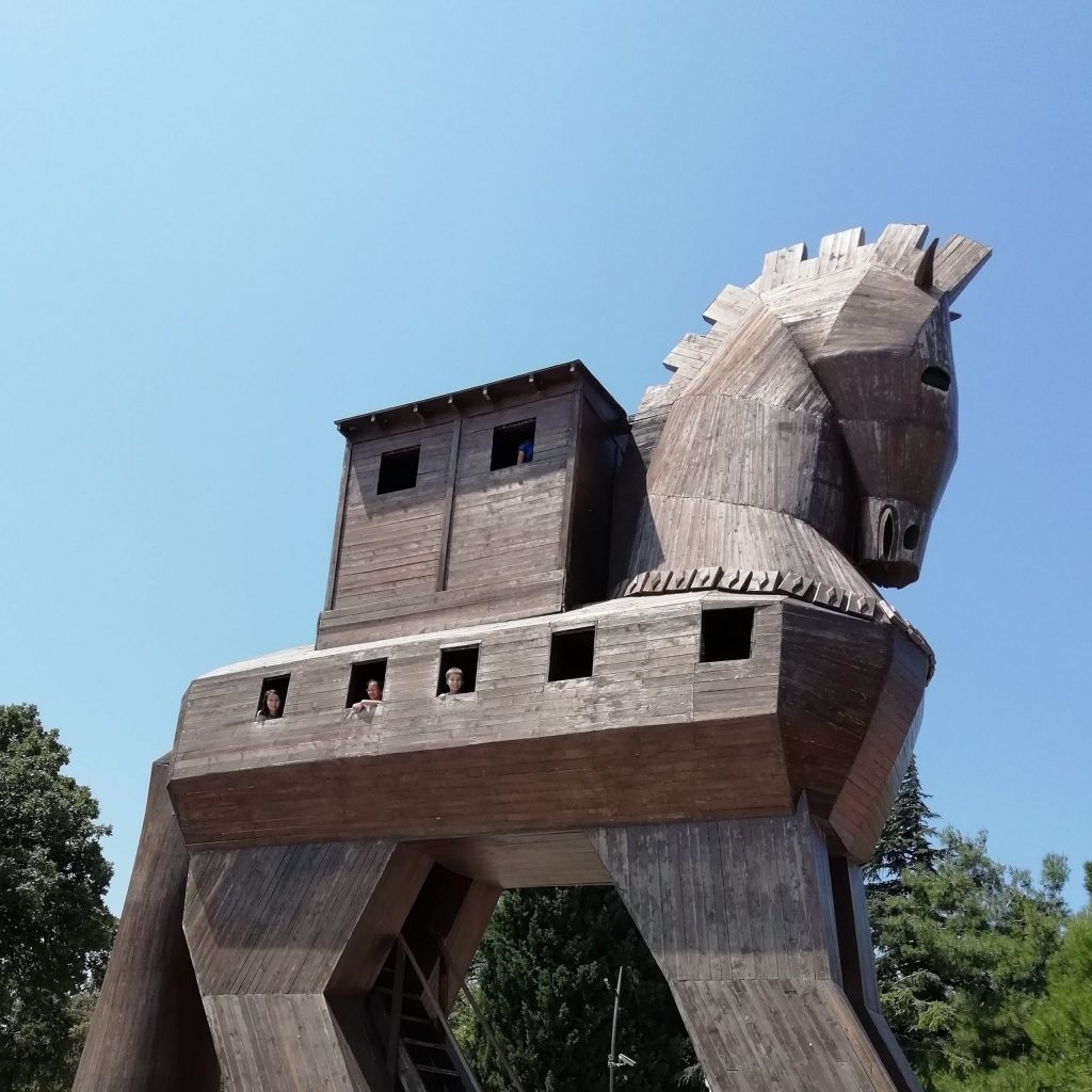 Wooden replica of the Wooden Horse of Troy at the entrance to the archaeological site; the head of the wooden horse is silhouetted against a pale blue sky.