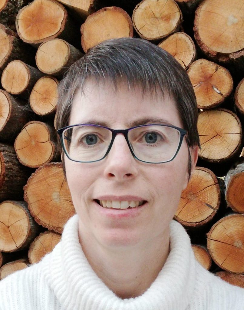 A woman with short brown hair and glasses stands in front of stacked logs